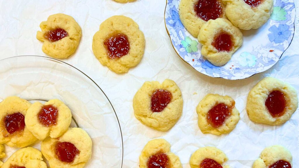 A collection of thumbprint cookies on a piece of parchment paper. There are some on a blue floral plate and some on a glass plate.