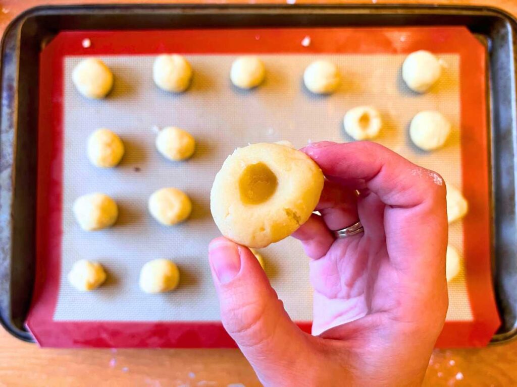 A woman is holding up an unbaked thumbprint cookie. 