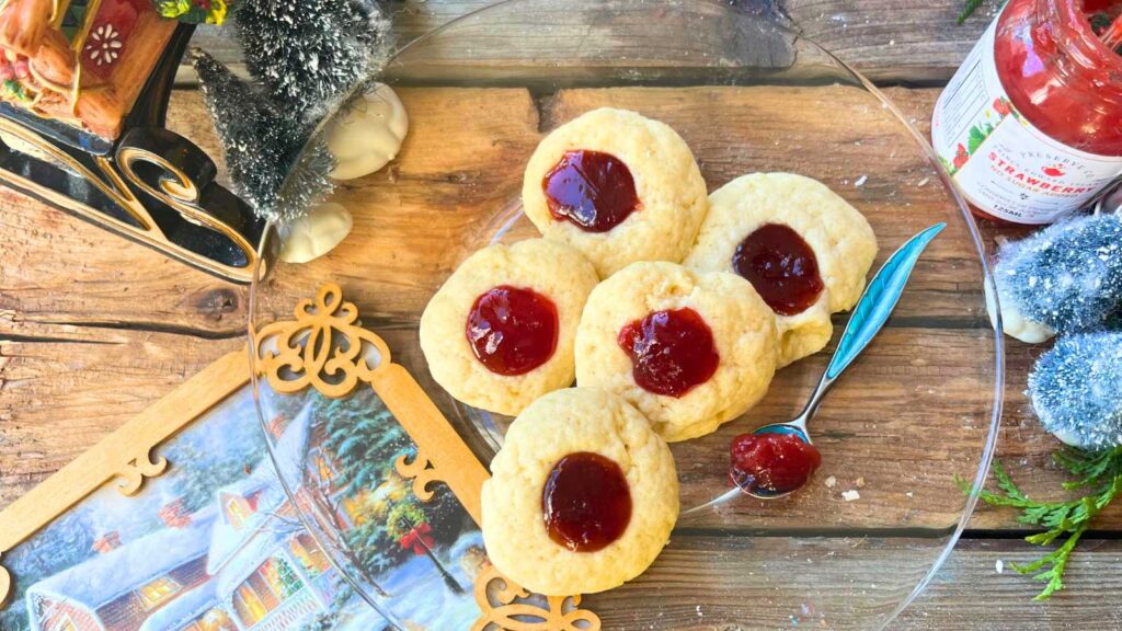 Five thumbprint cookies on a glass plate. There is jam in the centre of each cookie.