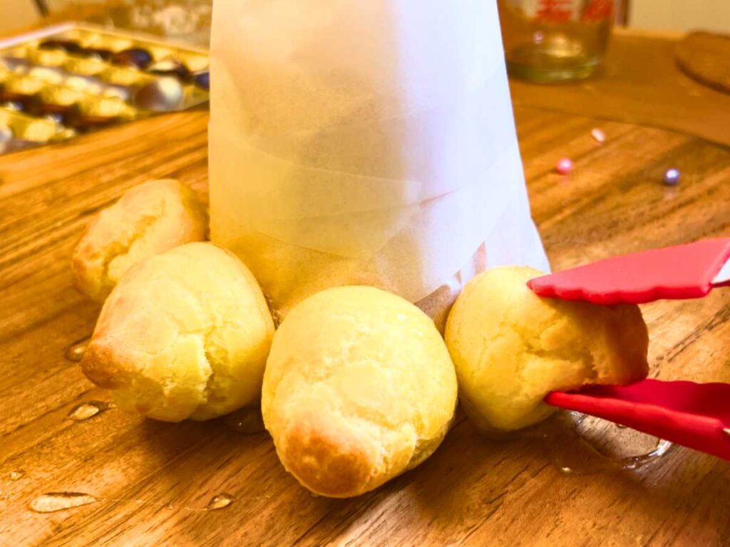 A woman is using red tongs to stick profiteroles onto a cone form.