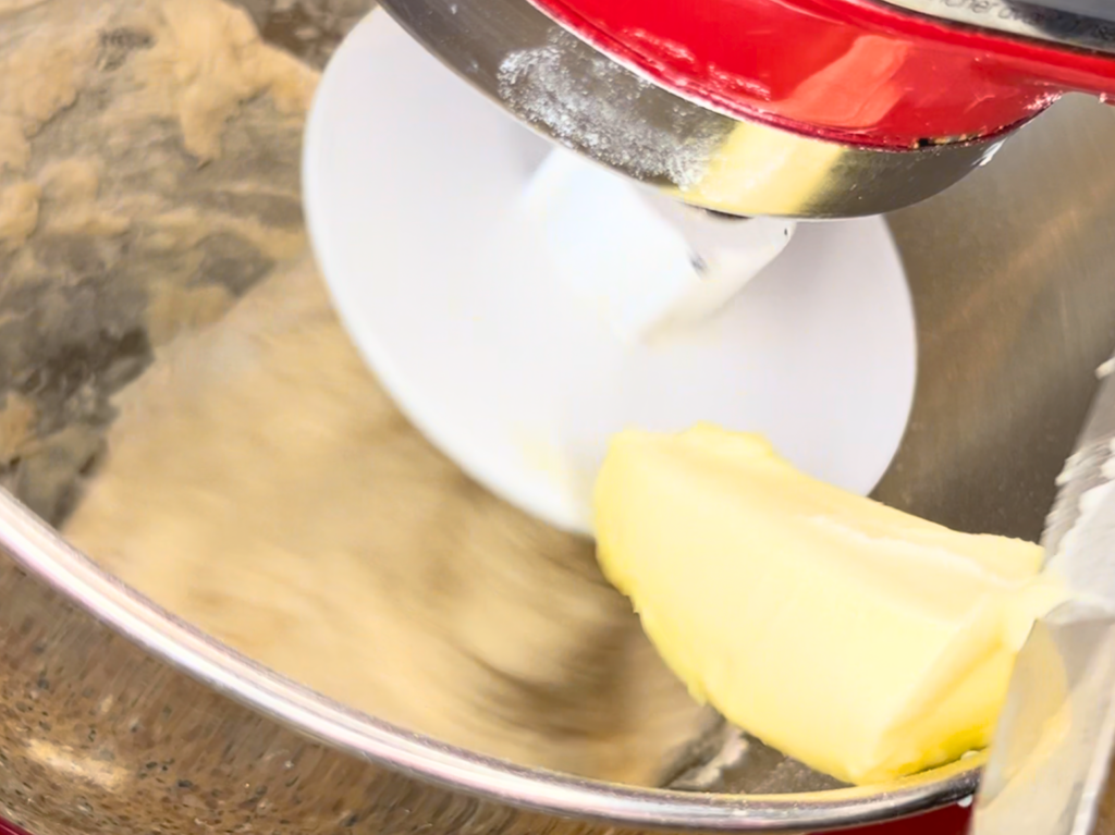 Dough being mixed in a stand mixer with a dough hook attachment. Butter is being added to the mixture.