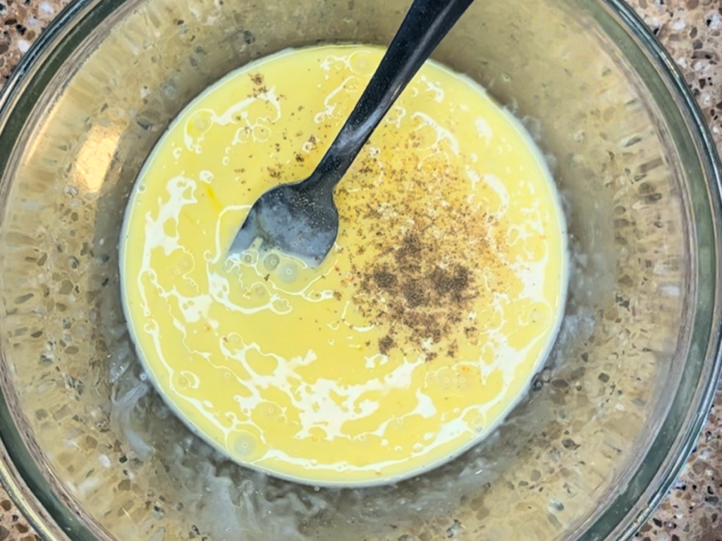 A glass bowl with a mixture of eggs, cream, and salt and pepper. There is a fork in the bowl.
