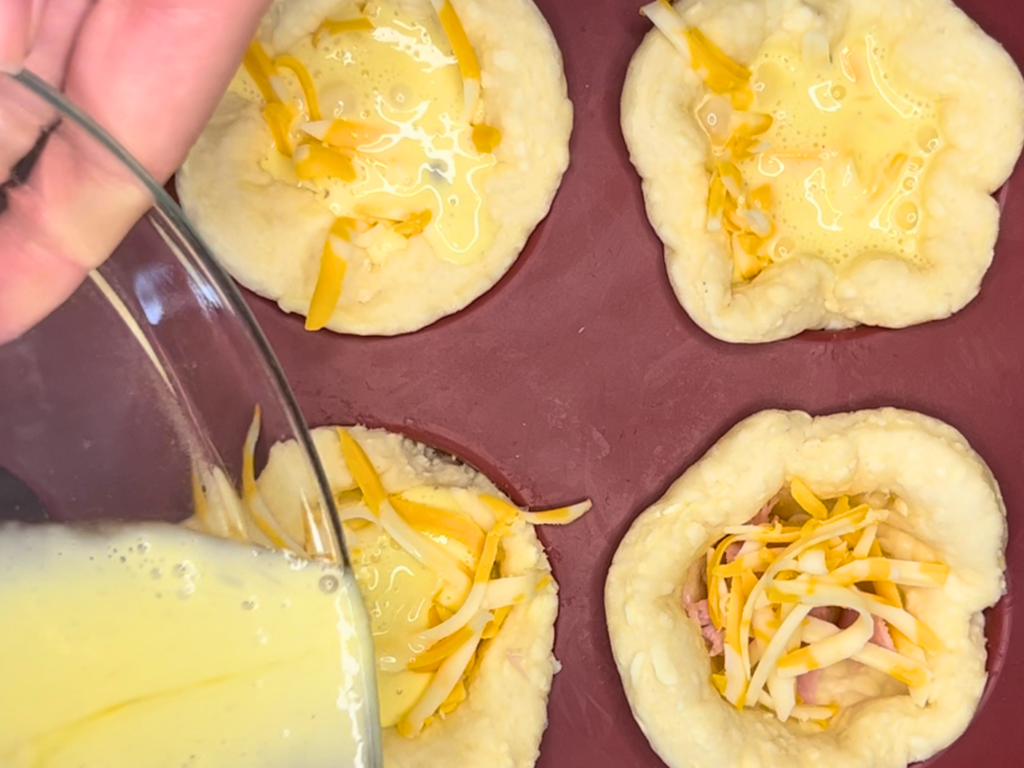 Woman pouring the egg mixture into puff pastry lined muffin cups with ham and cheese in them.