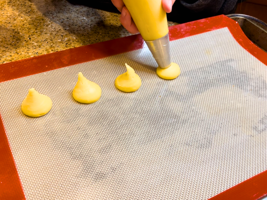 Woman piping pastry dough to make profiteroles onto a lined baking sheet.