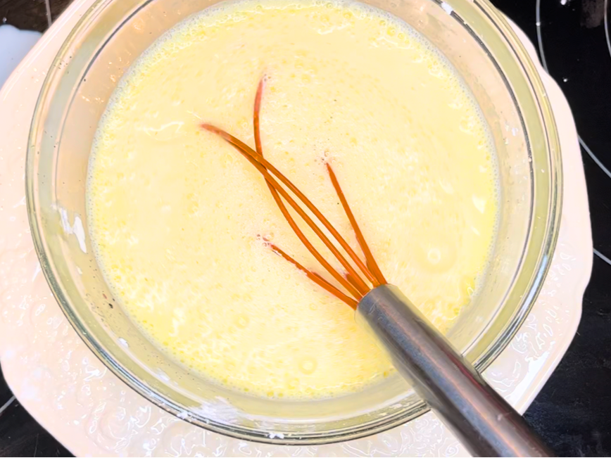 A glass bowl with a tempered egg and milk mixture. There is a whisk in the bowl.
