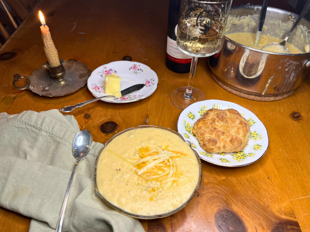 A table set for dinner. There is a bowl of potato soup in the foreground. Behind is a plate with a biscuit, and a glass of white wine. There is a lit candle and the full pot of potato soup is in the background. 