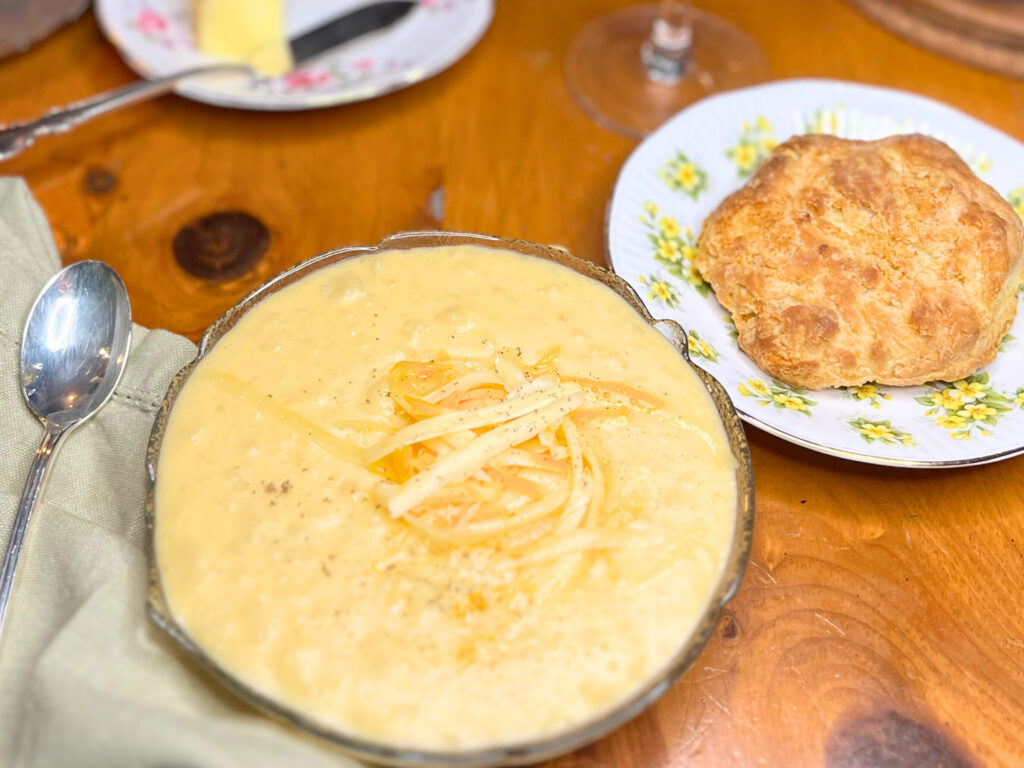 A glass bowl with potato soup in it. There is a sprinkle of grated cheese on top. A plate with a biscuit is in the background.