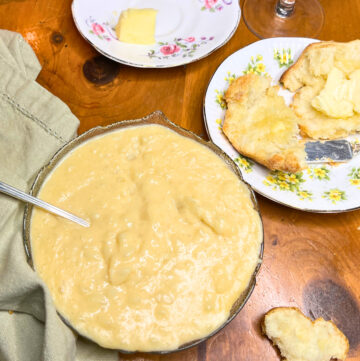 A glass bowl with potato soup. There is a spoon inside the bowl. A cut biscuit in the background with butter on it.