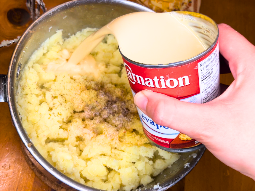 Woman pouring a can of evaporated milk over top of mashed potatoes. There is salt, pepper, and garlic powder on top of the potatoes.
