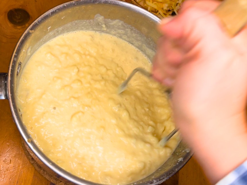 Woman stirring potato soup with a potato masher, inside of a metal sauce pot.