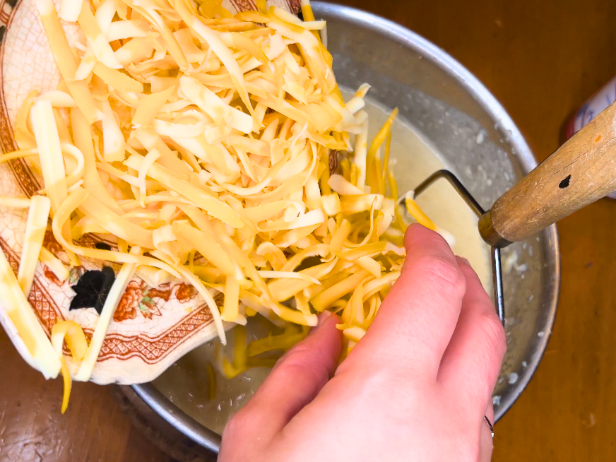 Woman adding grated cheese to a sauce pot of potato soup.