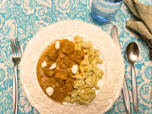 A white floral plate with chicken paprikash and nokedli on it. There is a place setting with cutlery, a glass of water and a napkin of to the side.