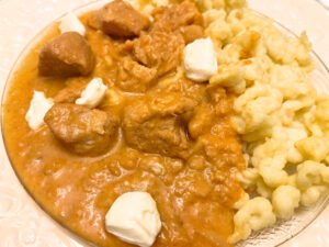 A white plate with chicken paprikash and spaetzle. There are some dollops of cream cheese on top.  