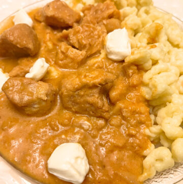 A white plate with chicken paprikash and spaetzle. There are some dollops of cream cheese on top.