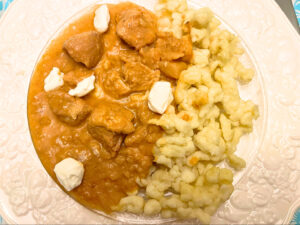 A white floral plate with spaetzle noodles and chicken paprikash. There is a few dollops of sour cream on top.