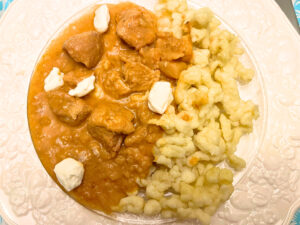 A white floral plate with chicken paprikash and spaetzle. There are some dollops of sour cream on top.