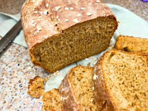 A loaf of brown bread with oats on top. There are slices of bread in the foreground.