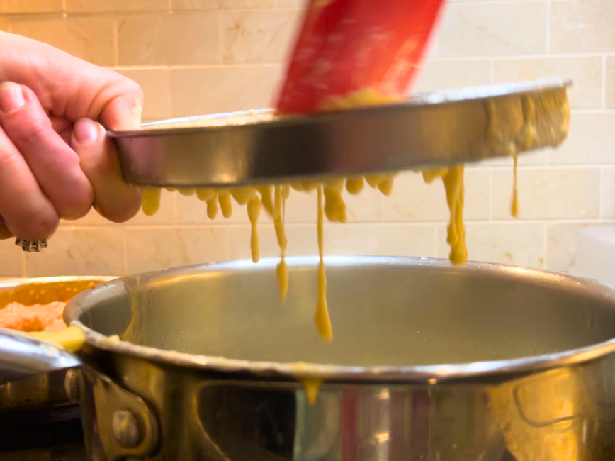 Woman pressing spaetzle batter through a cheese grater with a red spatula. The batter is falling into a pot of boiling water.