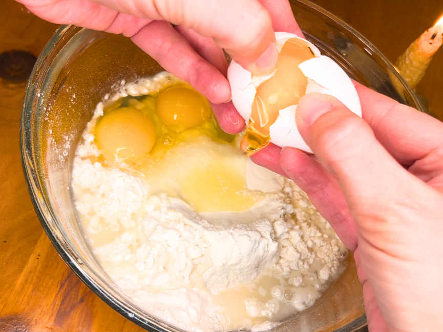 Woman cracking an egg into a large bowl with flour and two other cracked eggs.
