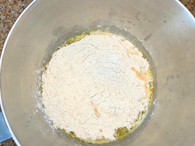 Whole wheat flour on top of a liquid mixture in a large metal bowl.