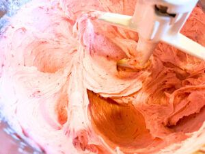 Light pink icing being mixed in a stand mixer fitted with the paddle attachment.