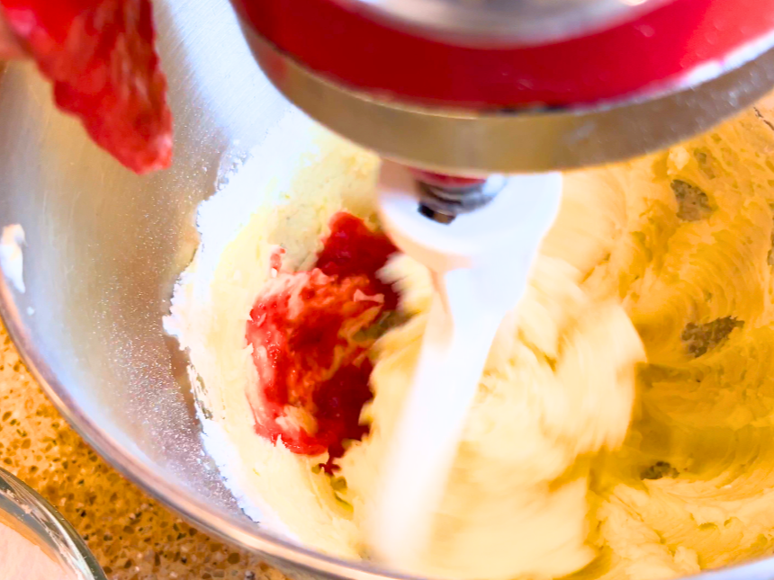 Strawberry sauce being added to the bowl of a stand mixer fitted with the paddle attachment. There is creamed butter and icing sugar in the bottom of the bowl.