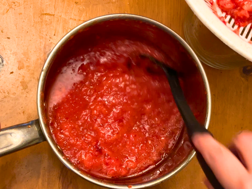 Woman stirring a pot of strawberry juice. There is a white strainer off to the side with mashed strawberries in it.