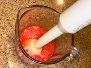 A glass measuring cup with pureed strawberries in it. There is an immersion blender in the cup as well.