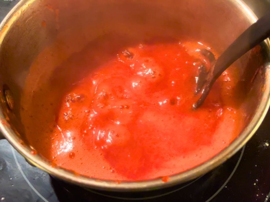 Strawberry juice in a metal sauce pot. The sauce is simmering and there is a black spoon in the pot.