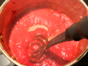 Woman stirring a pot of strawberry sauce. The sauce is thick. She is holding a black spoon.