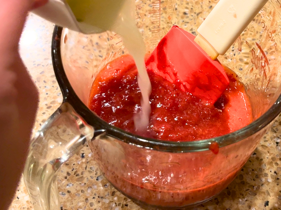 Woman pouring lemon juice into a glass measuring cup with thick strawberry sauce and some pureed strawberries. There is a red spatula in the glass as well.