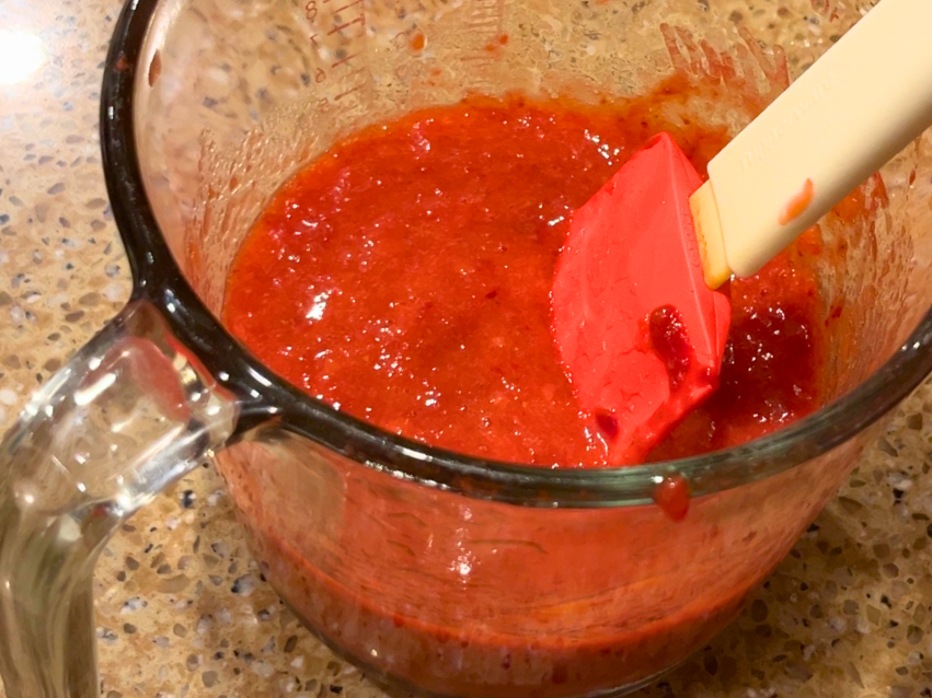 A glass measuring cup with strawberry sauce in it.  There is a red and white spatula in the glass as well.