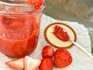 A jar of strawberry sauce with the lid off. A spoonful of sauce is resting on the lid. There are strawberries in the forground.