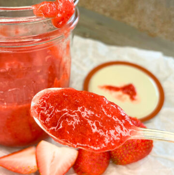 A jar of strawberry sauce with the lid off. There is a spoonful of the sauce in front and cut strawberries in the foreground.