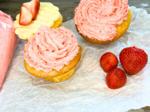 Three vanilla cupcakes sitting on some parchment paper. Two are decorated with pink icing and one with vanilla icing with cut strawberries on top. There are three strawberries in the foreground.