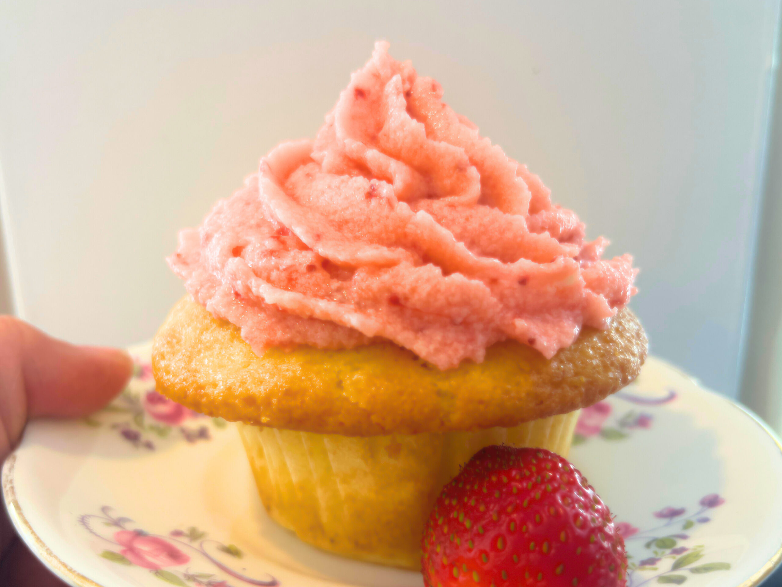 Woman holding a pink floral plate with a vanilla cupcake with pink icing on top. There is a strawberry on the plate as well.