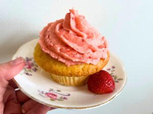 A vanilla cupcake with pink icing on top. There is a strawberry in front. They are sitting on a pink floral plate.