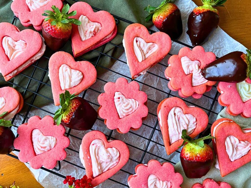 A collection of pink strawberry sandwich cookies on a wire rack. There are chocolate dipped strawberries.