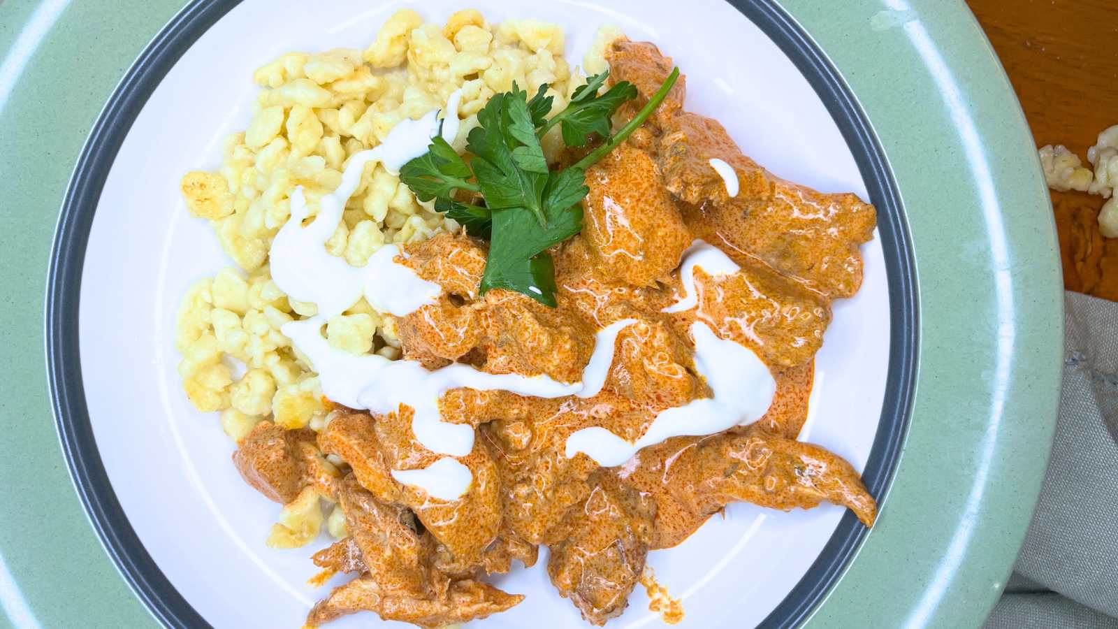 A white plate with a green rim with noodles and chicken paprikash garnished with sour cream and parsley.