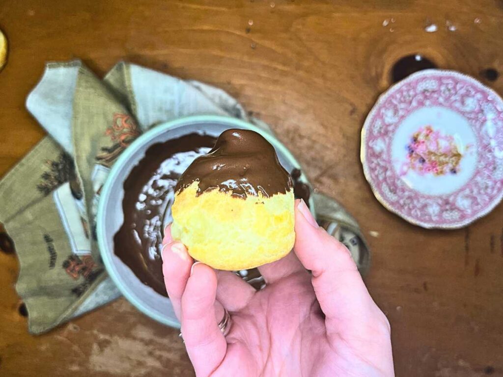 A woman is holding up a profiterole that has just been dipped in chocolate.