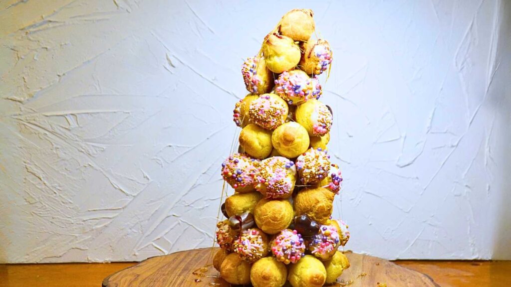 A croquembouche decorated with pink sprinkles on a wooden serving tray.