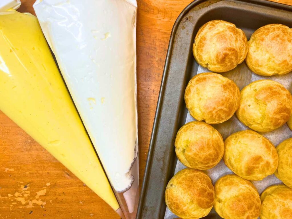 A tray full of profiteroles. There are two piping bags off to the side with a yellow and white cream inside.