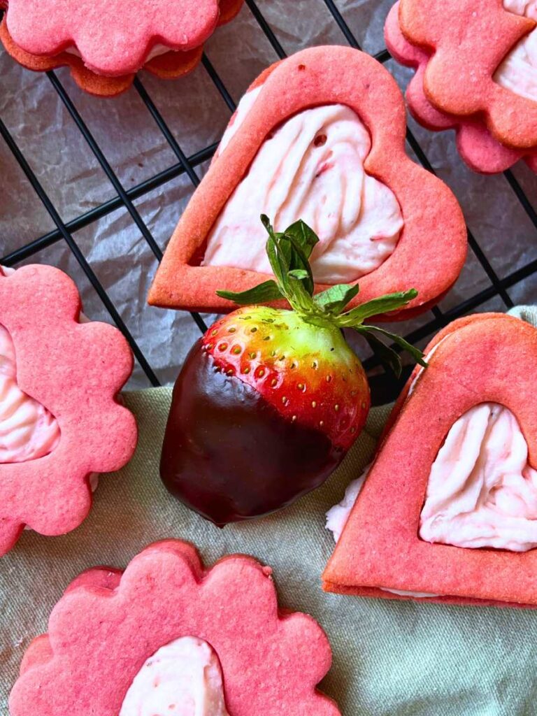 Heart shaped pink sandwich cookies with a chocolate dipped strawberry in the centre.