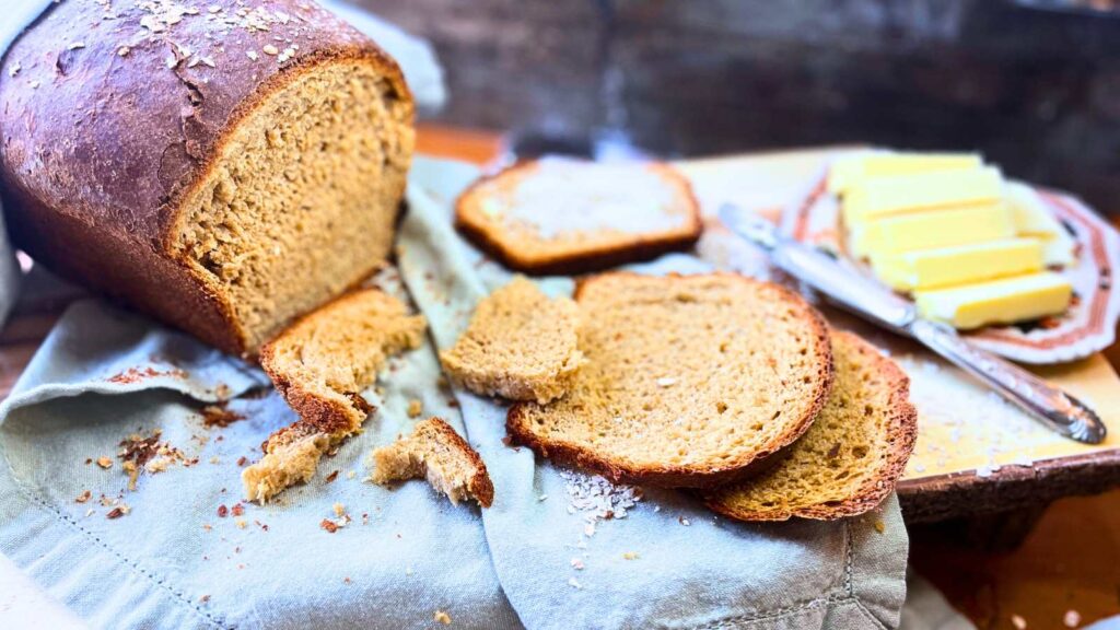 A sliced loaf of brown molasses bread. one slice has butter spread on it.