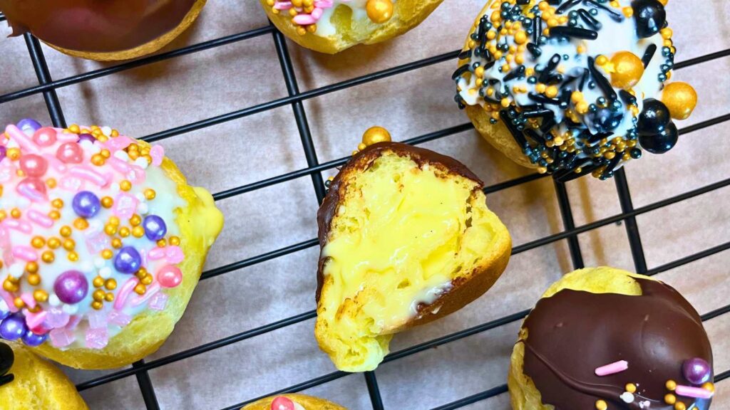 Profiteroles on a wire rack. One is split in half to show the pastry cream inside.