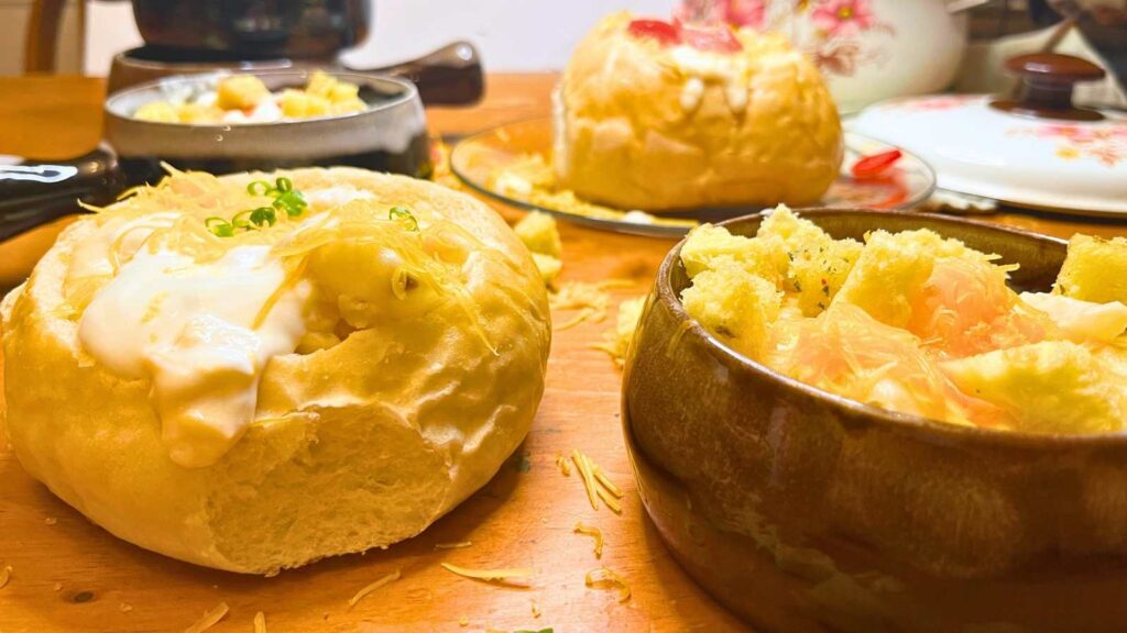 A side view of potato. One is in a bread bowl and one in a brown bowl. They are topped with croutons, cheese and chives.