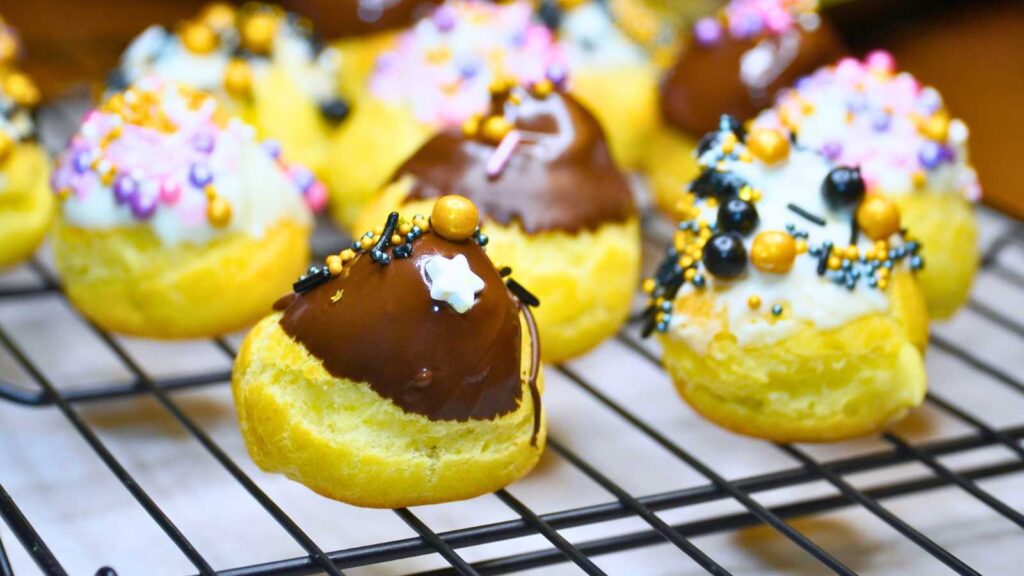 A group of profiteroles in a wire rack. Some coated in white chocolate and some in semi-sweet chocolate. All have sprinkles.