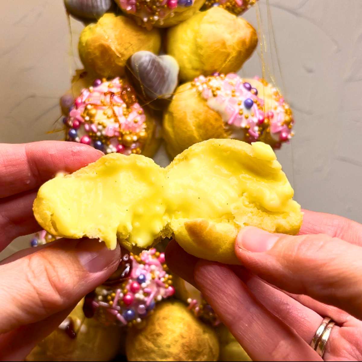 A woman is holding a profiterole showing the inside filling. There is a croquembouche in the background.