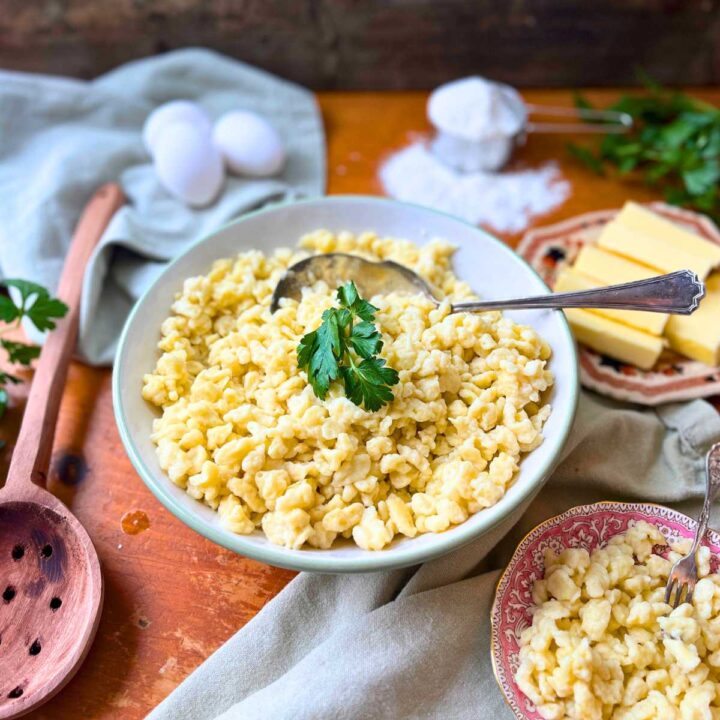 A large white bowl full of spaetzle noodles. There are eggs, flour, and butter in the background.