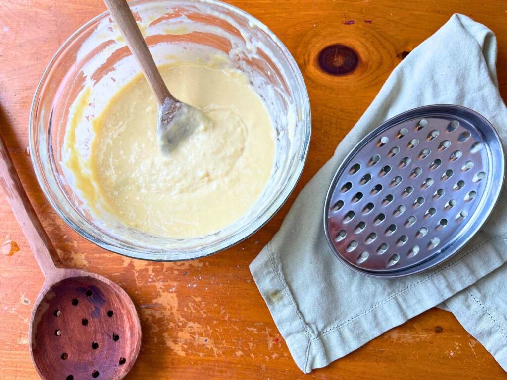 A glass bowl with spaetzle batter. There is a cheese grater and a wooden slotted spoon beside.